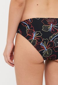 Bas de bikini noir avec un motif floral coloré en rouge, bleu et jaune. Il dispose d'une taille mi-haute et d'une texture lisse. Logo de la marque visible.