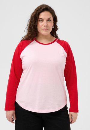 Femme aux cheveux bruns bouclés portant une chemise blanche à manches longues avec des manches rouges et un pantalon noir, debout devant un fond clair.