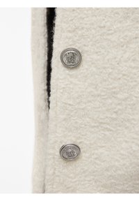 Gros plan sur un tissu à texture crème avec deux boutons décoratifs en argent ornés d'emblèmes de couronne et de bouclier.