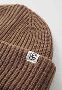 Chapeau en tricot marron avec une texture côtelée et un revers replié, arborant une étiquette blanche avec un logo noir sur le devant.