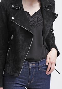 Czarna zamszowa kurtka motocyklowa z srebrnymi suwakami, noszona na czarnym topie z teksturowanym, falowanym wzorem, w zestawie z ciemnoniebieskimi dżinsami.