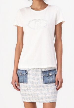 Mujer con una camiseta blanca de manga corta con un logo brillante y una falda de tweed blanca y azul con bolsillos de solapa vaqueros.