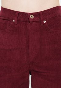 Pantalon en velours côtelé bordeaux en gros plan montrant la ceinture, le bouton avant, les passants de ceinture et les poches avant sur une taille à peau claire.