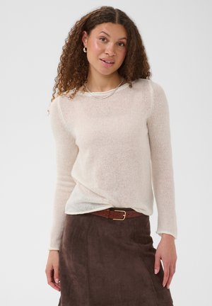 Femme debout et souriante, portant un pull léger à manches longues, une jupe marron avec ceinture, des boucles d’oreilles créoles et un collier sur fond uni.