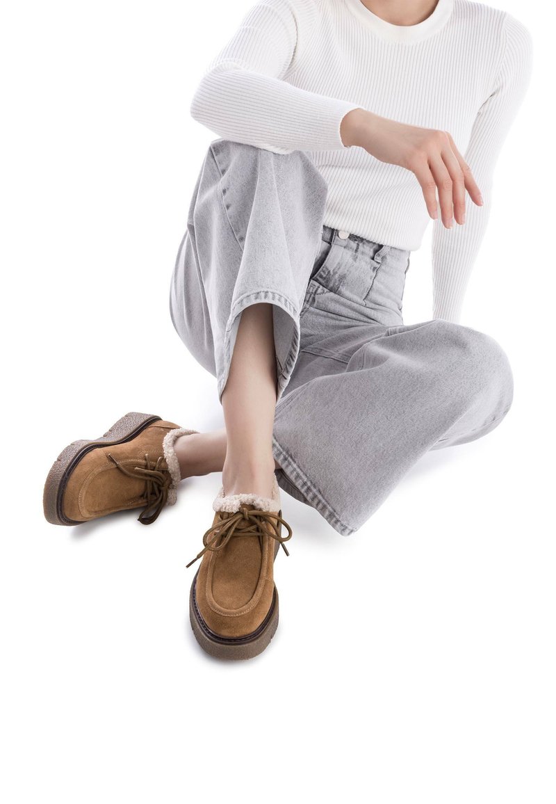 Zapatos de ante marrón con un cuello de piel suave y forrado, diseño con cordones y suela gruesa, combinados con jeans de pernera ancha grises y una blusa blanca de canalé.