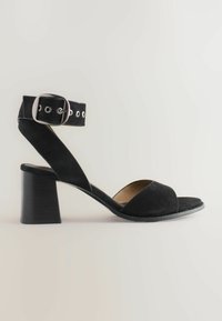 Black suede high-heeled sandal with an ankle strap and buckle, featuring an open-toe design and a chunky heel, against a light background.