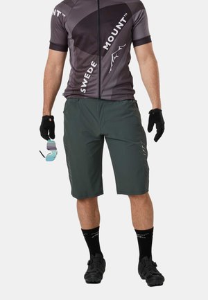Männlicher Radfahrer trägt ein graues und schwarzes Sweden Mount Radtrikot, dunkelgrüne Shorts, schwarze Handschuhe, Socken und Schuhe, hält eine Sonnenbrille in der linken Hand.