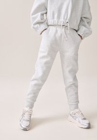 Helles graues Sweatshirt mit elastischen Bündchen an den Ärmeln und am Saum, kombiniert mit passenden Jogginghosen mit Taschen und gerippten Bündchen. Silberne Laufschuhe.