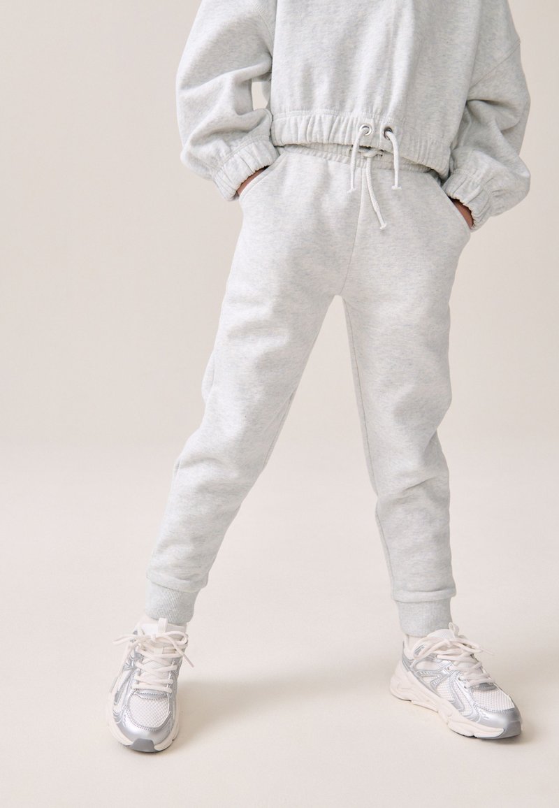 Helles graues Sweatshirt mit elastischen Bündchen an den Ärmeln und am Saum, kombiniert mit passenden Jogginghosen mit Taschen und gerippten Bündchen. Silberne Laufschuhe.