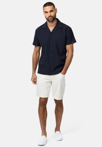 Camisa de botones azul marino de manga corta con cuello de muesca, combinada con pantalones cortos claros enrollados. El modelo lleva zapatos slip-on blancos.