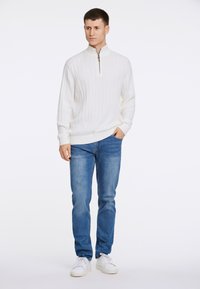 Weißer Kabelstrick-Pullover mit hohem Kragen und Halb-Reißverschluss, kombiniert mit blauen Jeans und weißen Sneakers. Einfaches, klassisches Design mit strukturiertem Stoff.