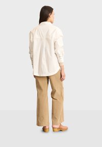 Lichtbeige oversized knoopshirt met drop schouders en een gebogen zoom, gecombineerd met wijde corduroybroek in een tan kleur. Bruine gehakt clogs.