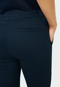 Mörkblå, figurnära byxor med ett texturerat rutmönster, med en bakficka och en smal silhuett. Materialet verkar slätt.