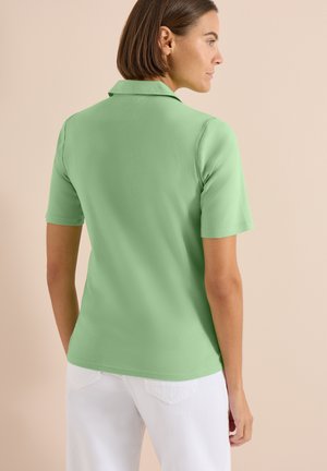 Femme aux cheveux courts et bruns, portant un polo vert clair à manches courtes et un pantalon blanc, debout devant un fond beige.