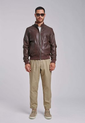 Mężczyzna w ciemnobrązowej skórzanej kurtce, beżowych spodniach, beżowych butach i okularach przeciwsłonecznych, stojący na tle jednolitego jasnoszarego tła.