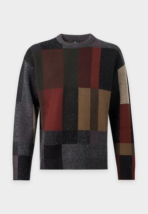 Jeune homme debout, les mains dans les poches, portant un pull patchwork multicolore, pantalon gris et chaussures marron sur fond blanc.