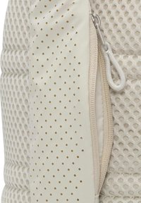 Vitt, perforerat tyg med guldbollar har en dragkedja längs sidan. Texturen är slät, med en vadderad, quiltad design.