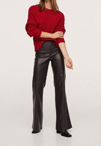 Maglione rosso lavorato a maglia con motivi a trecce; pantaloni svasati neri in ecopelle; il modello indossa stivaletti neri alla caviglia; sfondo neutro.