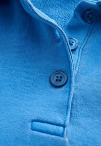 Lichtblauwe poloshirtstof met een zachte textuur, voorzien van een knoopsluiting met donkerblauwe knopen en een klein borstzakje.