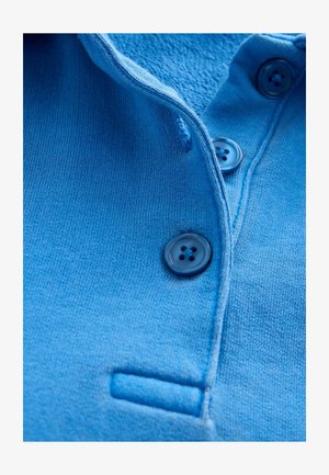 Tessuto della polo azzurro chiaro con una consistenza morbida, dotata di una chiusura con bottoni di colore blu scuro e un piccolo taschino sul petto.