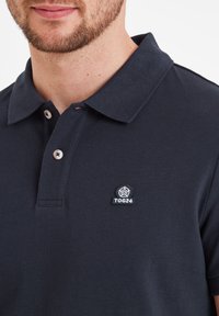 Navy polo shirt in textured fabric with a classic collar. Features a left chest logo patch and three buttons down the placket.