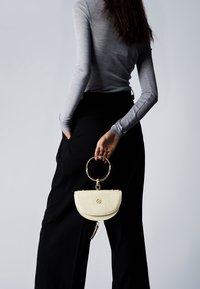 Borsa a mezzaluna circolare in ecopelle testurizzata color crema con manico circolare in tonalità oro, dotata di logo e dettagli con nappine decorative.