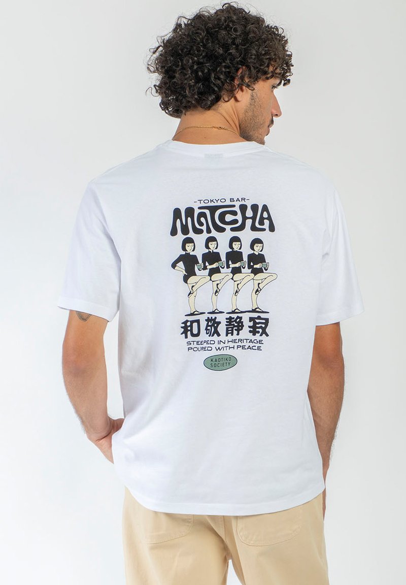 Άνδρας με σγουρά μαλλιά που φοράει λευκό t-shirt με γραφικό Tokyo Bar Matcha που δείχνει τέσσερις γυναίκες να κρατούν φλιτζάνια σε σειρά.