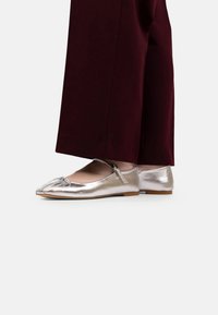 Balerini metalizate argintii cu vârful rotund, având o curea și plissare subtilă pe partea superioară. Purtați cu pantaloni largi bordeaux.