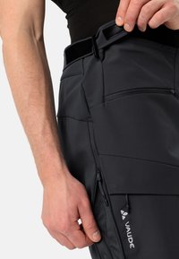 Pantalons extérieurs noirs avec poche zippée, ceinture ajustable et tissu texturé ; présente un logo de marque et plusieurs coutures pour le design.