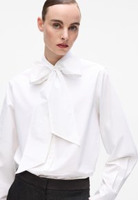 White blouse with a large bow tie at the collar, featuring long sleeves and a button-up closure. The fabric appears smooth and crisp.