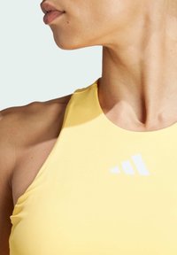 Top sportivo giallo con una texture liscia e colletto alto. Presenta un logo bianco sul lato sinistro e un design aderente che si adatta al corpo.