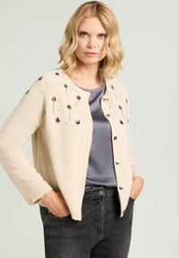 Beige Cardigan mit dekorativen Pailletten und Perlen, Rundhalsausschnitt, Knopfverschluss, gerippte Bündchen, getragen über einem grauen Satin-Top und dunkler Jeans.