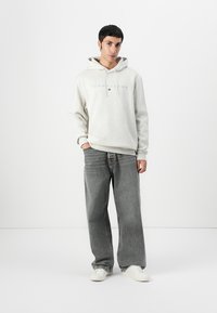 Lys grå hettegenser med snorhette og Tommy Jeans-logo, kombinert med løse grå jeans og hvite sneakers. Glatt tekstur.