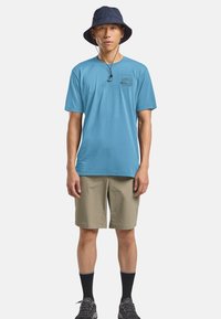 Lichtblauw T-shirt met korte mouwen met een grafisch ontwerp aan de voorkant, gecombineerd met kaki shorts, zwarte sokken en een donkerblauwe bucket hat.