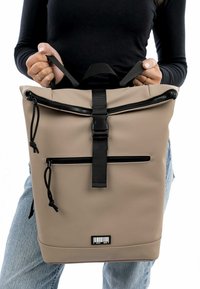 Zaino beige realizzato in materiale impermeabile, dotato di cerniere nere, una tracolla regolabile, una tasca frontale e un dettaglio con codice a barre.