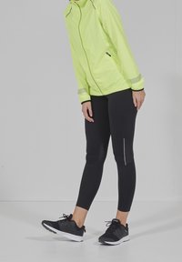 Osoba v neonově zelené bundě, černých legínách s reflexními detaily a černých sportovních botách stojí na bílé podlaze na pozadí šedé stěny.