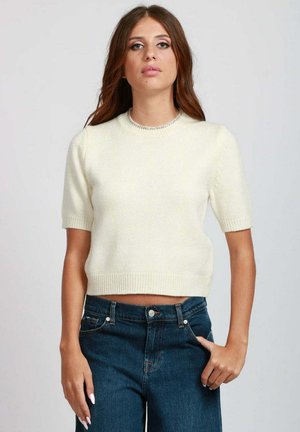 Maglione corto di colore crema con maniche corte, scollatura rotonda e bordi a costine. Tessuto liscio, indossato con jeans blu scuro.