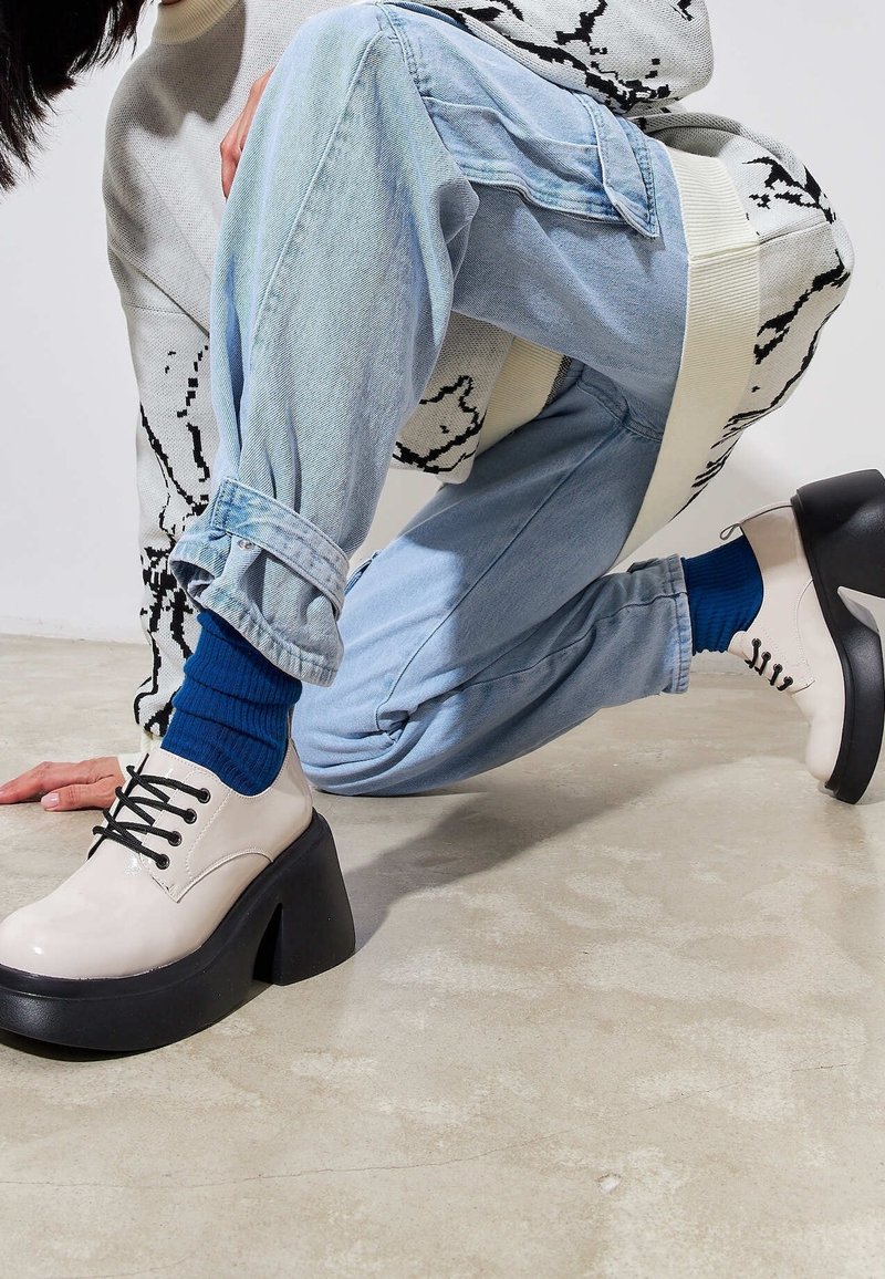 Personne à genoux en jean bleu clair avec revers roulés, pull crème avec motif noir, chaussettes bleues et chaussures compensées beiges à talons larges.