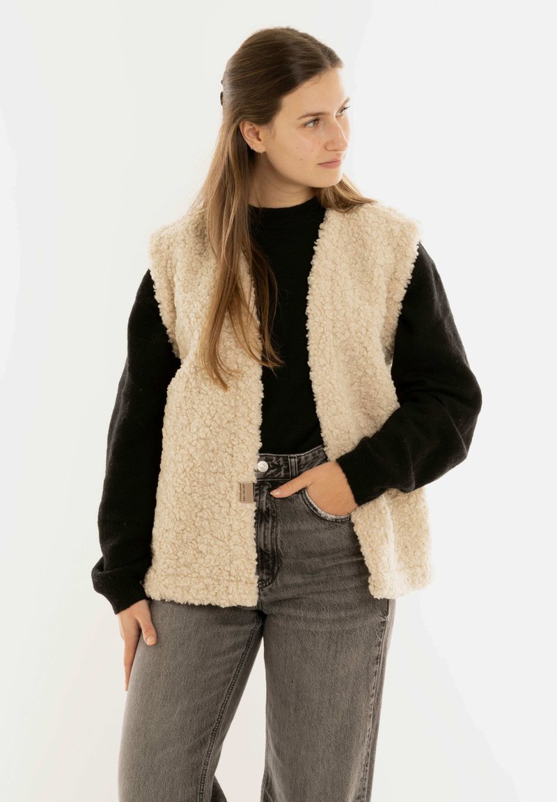 Beige Kunstfellweste mit offener Vorderseite, getragen über einem schwarzen Pullover und grauen hoch geschnittenen Jeans. Die Weste hat eine strukturierte Oberfläche.