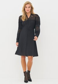 Schwarzes Kleid mit Spitzenapplikationen an den langen Ärmeln, einem V-Ausschnitt und einem Bindegürtel in der Taille. Der Stoff ist glatt und fällt knielang.