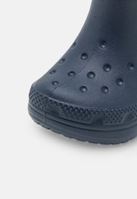 Gros plan sur un bout de botte en caoutchouc bleu foncé avec des bosselures circulaires surélevées et un bord de semelle texturé sur fond blanc.