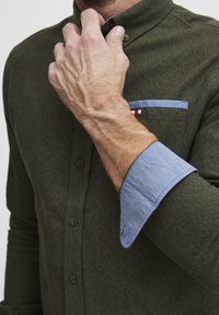 Mørkegrønn skjorte med strukturert stoff, knapp-til-ned krage og kontrasterende lyseblå mansjett. Har en brystlomme med stripet detalj.