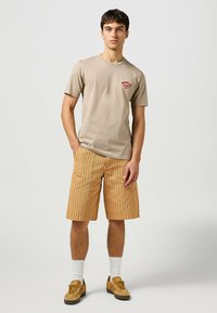 Beige bomulls t-shirt med röd logotyp på bröstet, matchad med orange randiga shorts. Modellen har på sig vita strumpor och bruna sneakers, med avslappnade armar.