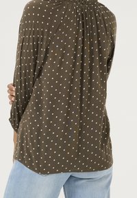 Blouse vert olive à manches longues avec des pois blancs, dotée d'un col froncé et d'une coupe ample. L'ourlet est légèrement plus long à l'arrière.