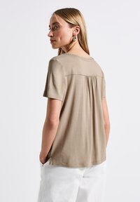 Krémová košile s krátkým rukávem a kulatým výstřihem, s nařaseným detailem na zádech a malými dekorativními stehy kolem límce. Hladká textura látky.