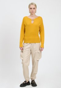 Gele gebreide trui met V-hals en een gouden sluiting, gecombineerd met beige neplederen cargobroek met zijzakken en elastische boorden.