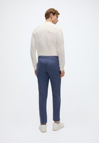 Μακρυμάνικο πουκάμισο σε ανοιχτό κρεμ χρώμα και σκούρο μπλε παντελόνι. Το παντελόνι έχει ίσια κοπή, ορατές τσέπες και φοριέται με ανοιχτόχρωμα αθλητικά παπούτσια.