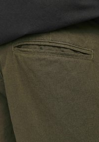 Pantalones verde oliva con una superficie texturizada, que presentan un bolsillo trasero horizontal con un borde cosido, parcialmente cubierto por una parte superior oscura.