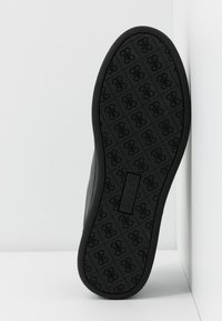 Semelle de chaussure noire avec un motif répété de logo entrelacé et une étiquette de logo rectangulaire au centre.