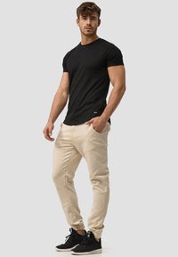 Camiseta negra ajustada, pantalones beige tipo taper con detalles de costura distintivos, combinados con zapatillas deportivas negras, sobre un fondo neutro.